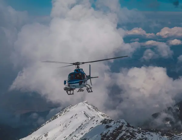 Ab Dezember 2026: glockna – das wohl coolste Hotel in Fusch Hubschrauber fliegt über schneebedeckten Berg vor wolkigem Himmel